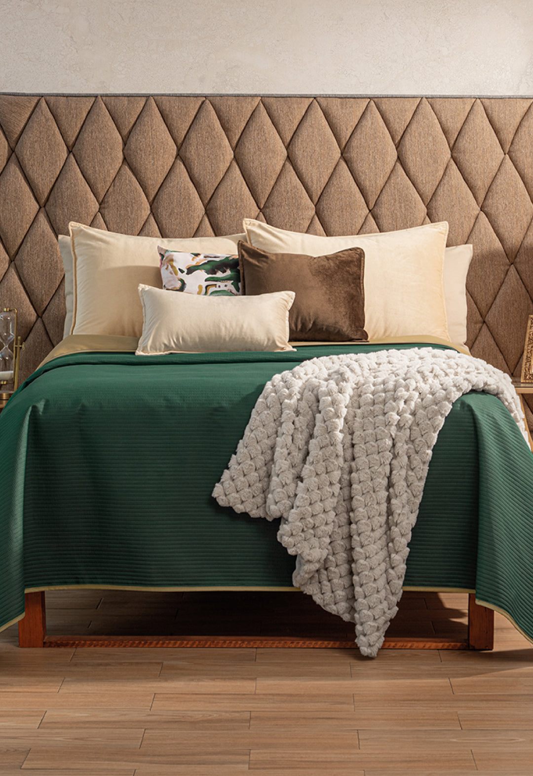 A neatly made bed showcases the Juego de Edredón Ligero Honor by Intima Hogar in green, accented with beige and brown pillows, a patterned cushion, and a textured white edredón capitonado. The diamond-patterned headboard and wooden floor complete the look.