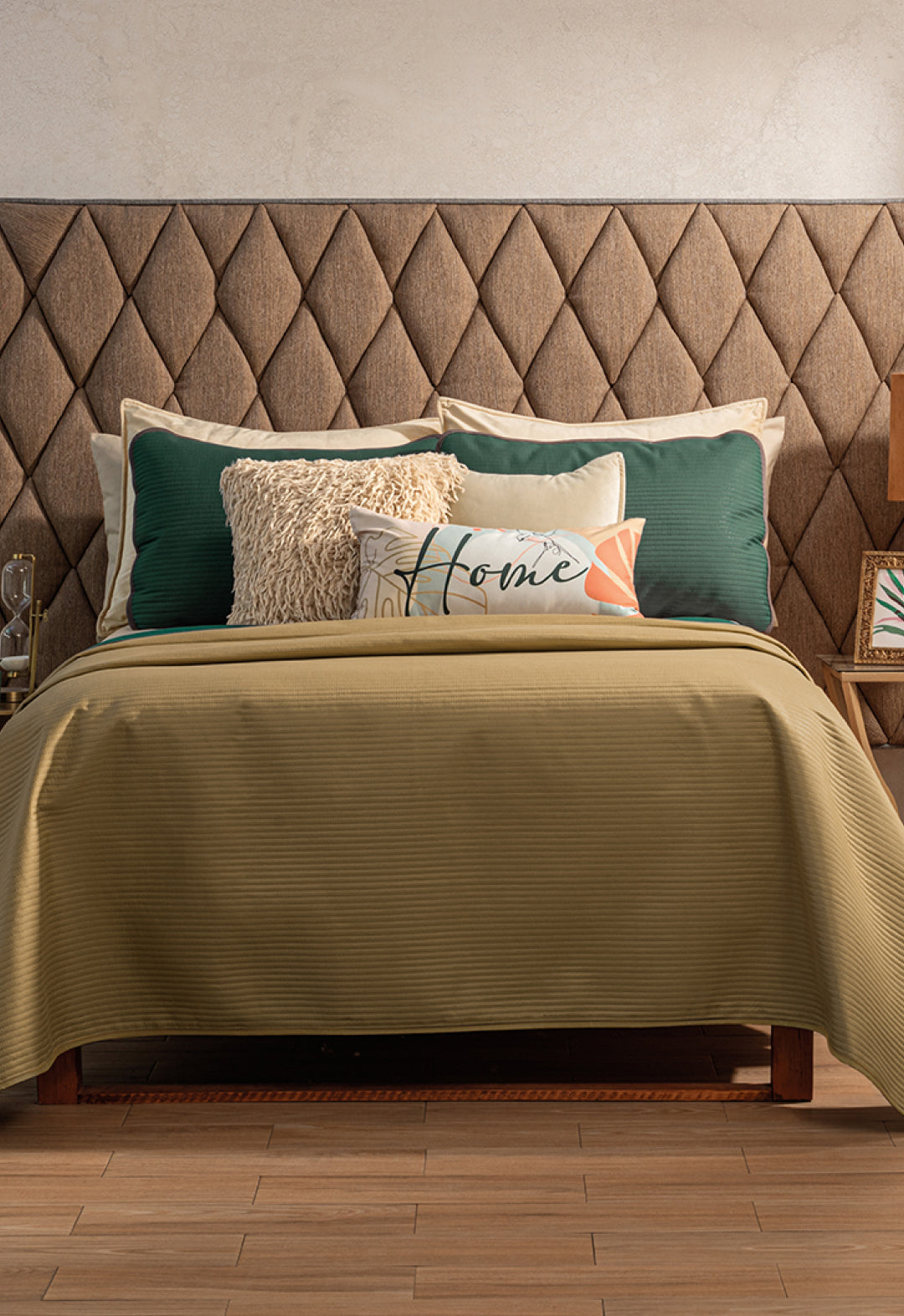 A bed featuring the Juego de Edredón Ligero Honor by Intima Hogar in olive green, with decorative pillows—one reading "Home"—and a beige, diamond-patterned headboard. Small nightstands sit on either side.
