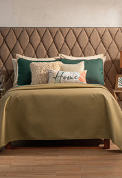 A bed featuring the Juego de Edredón Ligero Honor by Intima Hogar in olive green, with decorative pillows—one reading "Home"—and a beige, diamond-patterned headboard. Small nightstands sit on either side.