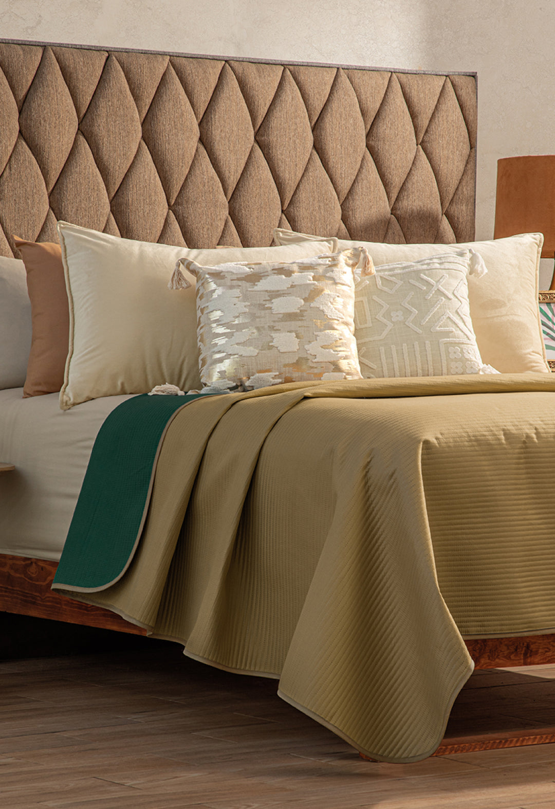 A neatly made bed featuring the Intima Hogar Juego de Edredón Ligero Honor, with a brown tufted headboard, gold and cream pillows, a green blanket at the edge, and set on a wooden floor.