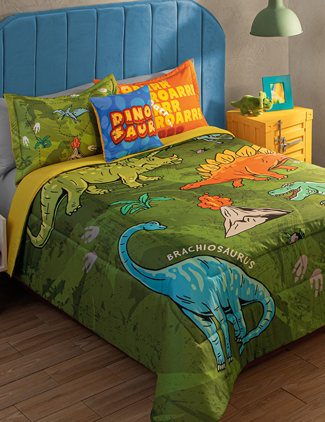 A cozy twin bed with a blue headboard features the Intima Hogar Juego de Edredón Jurásico, decorated with vibrant dinosaurs, plants, and footprints—ideal for una habitación infantil. A yellow nightstand holds a green lamp and toy dinosaur.