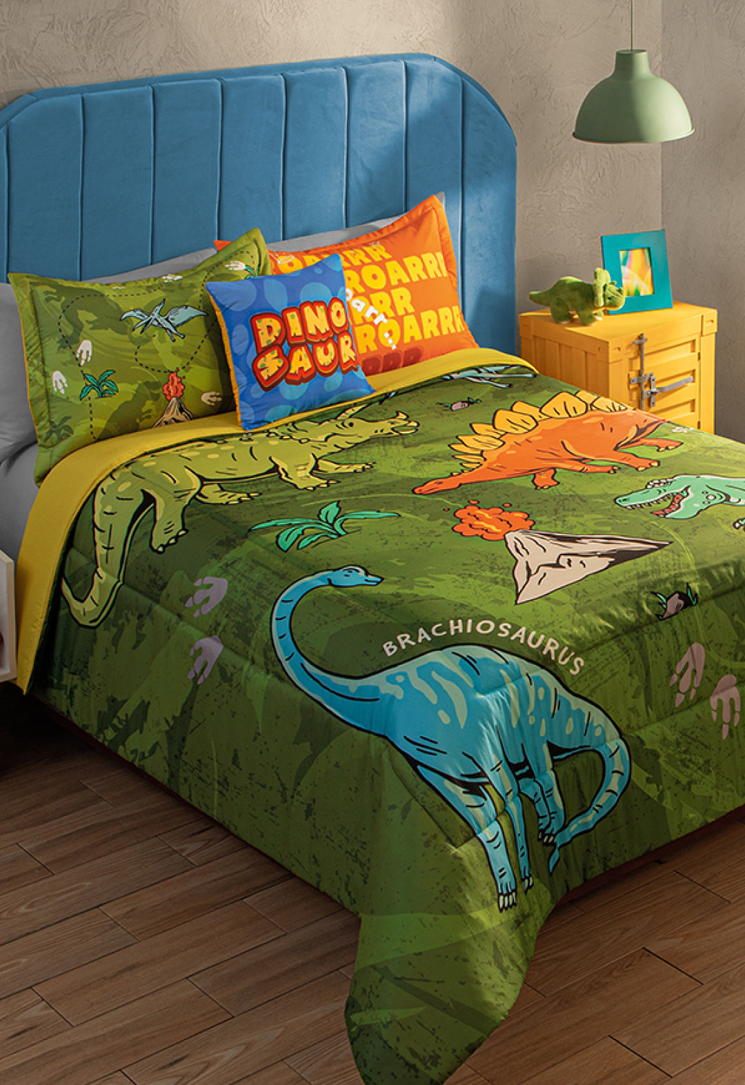 A cozy twin bed with a blue headboard features the Intima Hogar Juego de Edredón Jurásico, decorated with vibrant dinosaurs, plants, and footprints—ideal for una habitación infantil. A yellow nightstand holds a green lamp and toy dinosaur.