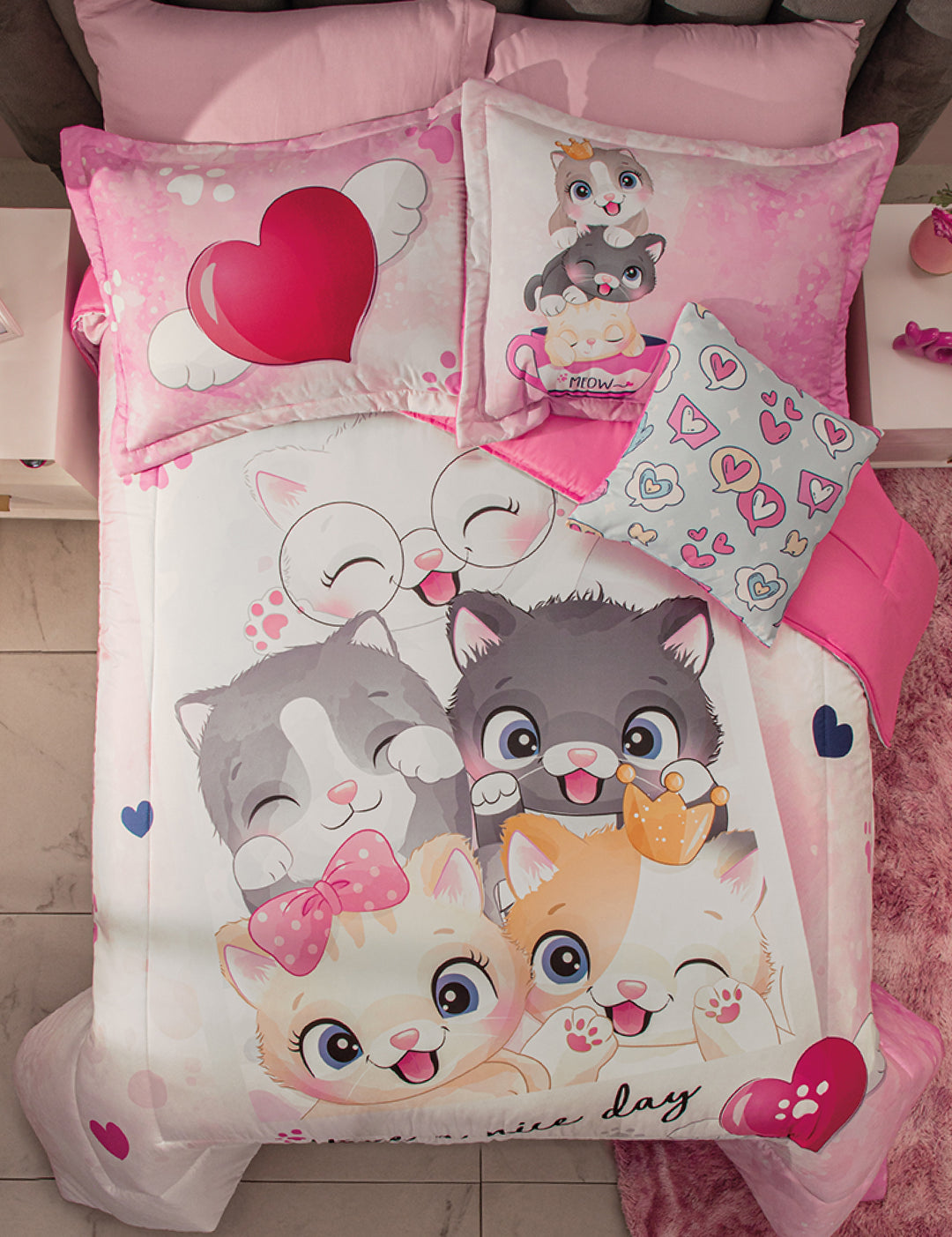 A neatly made bed in a cozy room features the Juego de Edredón Lindo Gatito by Intima Hogar, adorned with cute cartoon kittens, hearts in pink, white, and gray, plus a heart-print and a colorful heart throw pillow.