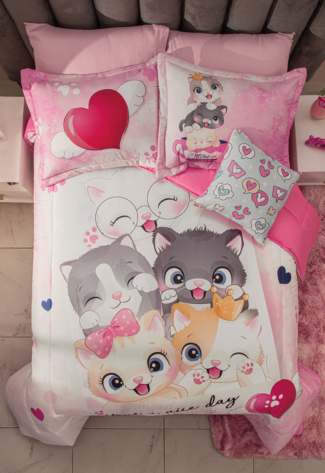 A neatly made bed in a cozy room features the Juego de Edredón Lindo Gatito by Intima Hogar, adorned with cute cartoon kittens, hearts in pink, white, and gray, plus a heart-print and a colorful heart throw pillow.