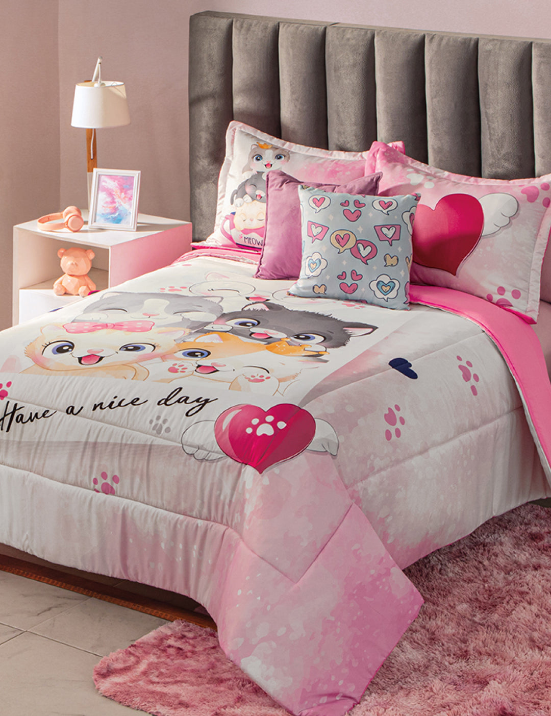 A neatly made bed features the Intima Hogar Juego de Edredón Lindo Gatito, matching pillows, and a heart cushion. The cozy room includes a gray headboard, pink rug, teddy bear, nightstand with lamp, and framed photo.