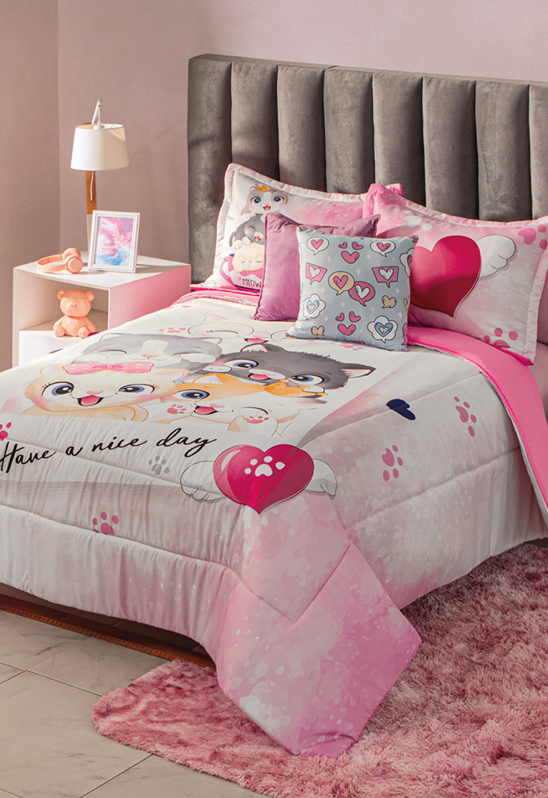 A neatly made bed features the Intima Hogar Juego de Edredón Lindo Gatito, matching pillows, and a heart cushion. The cozy room includes a gray headboard, pink rug, teddy bear, nightstand with lamp, and framed photo.