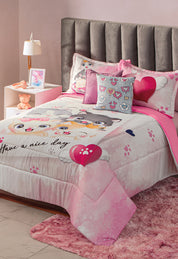 A neatly made bed features the Intima Hogar Juego de Edredón Lindo Gatito, matching pillows, and a heart cushion. The cozy room includes a gray headboard, pink rug, teddy bear, nightstand with lamp, and framed photo.
