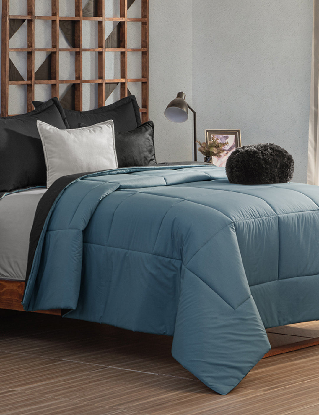 Neatly made bed with the Juego de Edredón Marco by Intima Hogar in blue, black and white pillows, and a round black cushion. A wooden geometric headboard and a bedside table with lamp and framed photo complete the scene.