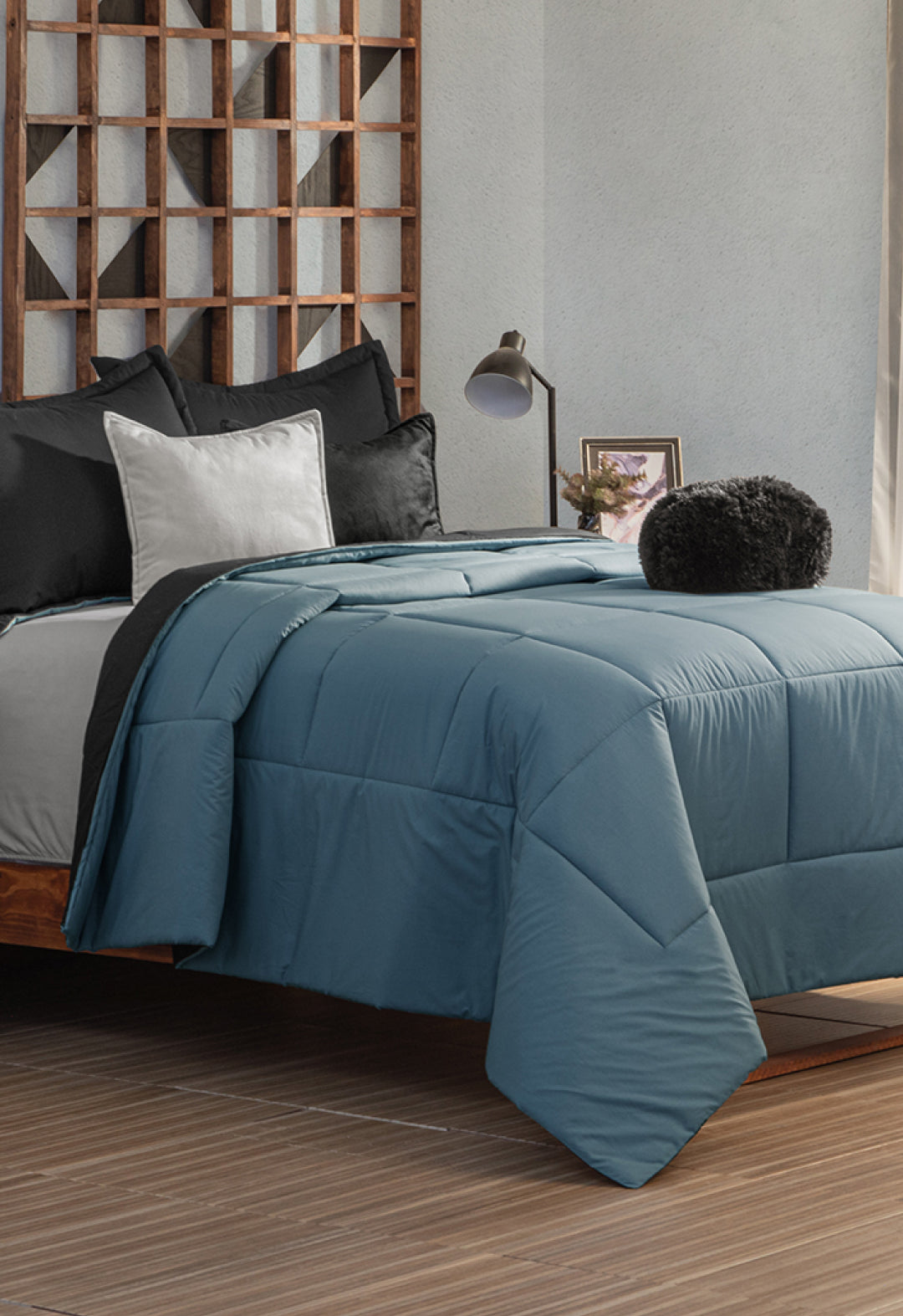 Neatly made bed with the Juego de Edredón Marco by Intima Hogar in blue, black and white pillows, and a round black cushion. A wooden geometric headboard and a bedside table with lamp and framed photo complete the scene.