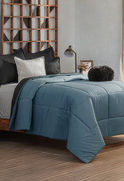 Neatly made bed with the Juego de Edredón Marco by Intima Hogar in blue, black and white pillows, and a round black cushion. A wooden geometric headboard and a bedside table with lamp and framed photo complete the scene.