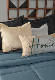 A neatly made bed with the Juego de Edredón Marco by Intima Hogar in blue microfiber, black and cream pillows, and a decorative "Home" pillow featuring abstract designs, all set against a wooden headboard with geometric accents for light warmth.