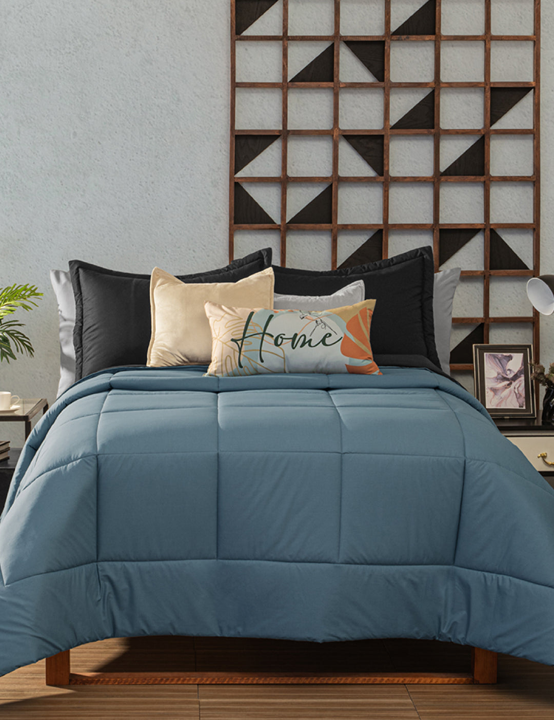 A neatly made bed with the Juego de Edredón Marco by Intima Hogar in blue, paired with black and beige pillows and a “Home” decorative pillow. The bed sits against a light wall adorned with geometric wooden art, flanked by nightstands and lamps.