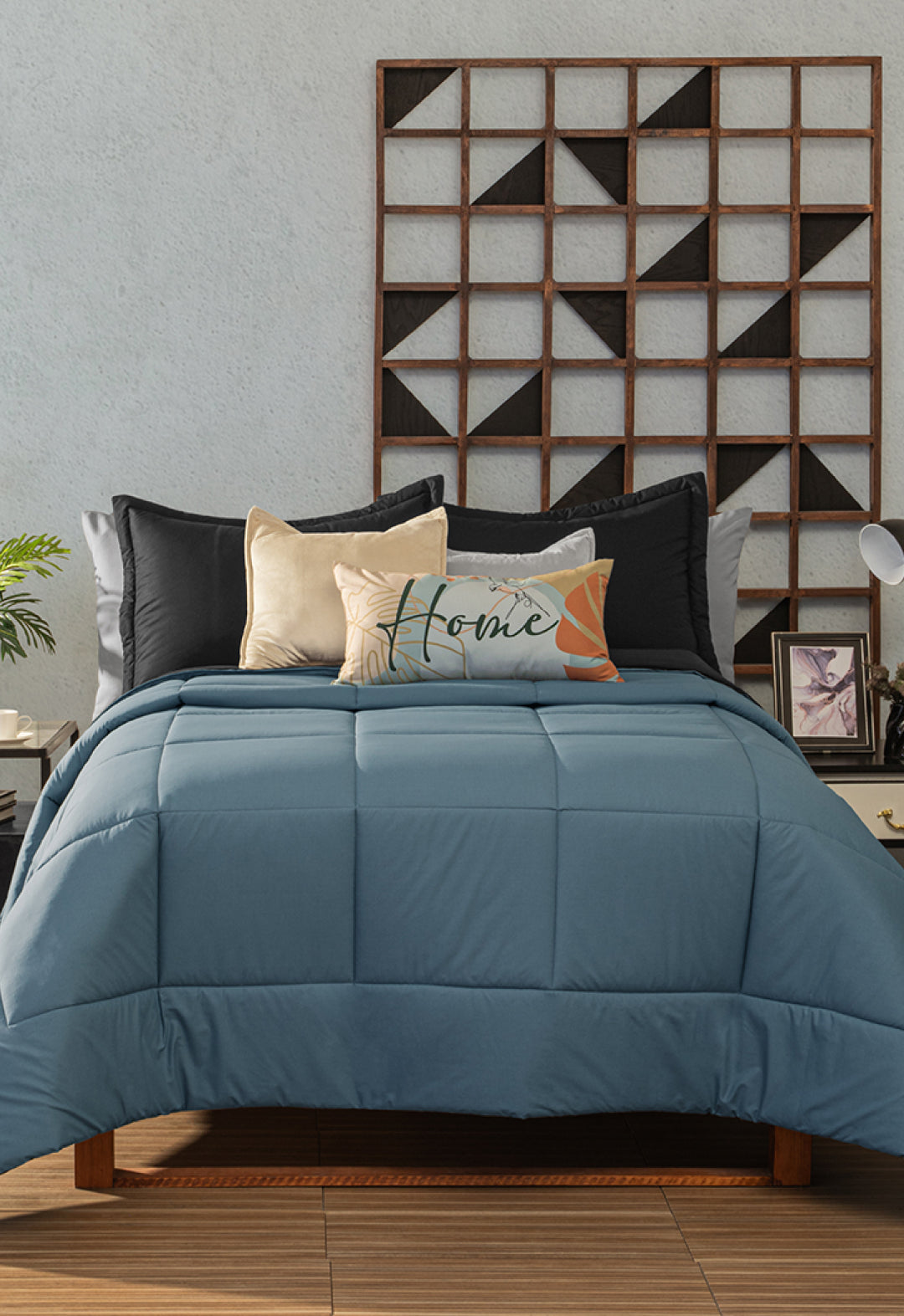 A neatly made bed with the Juego de Edredón Marco by Intima Hogar in blue, paired with black and beige pillows and a “Home” decorative pillow. The bed sits against a light wall adorned with geometric wooden art, flanked by nightstands and lamps.
