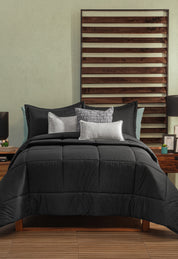 A neatly made bed features the Juego de Edredón Marco by Intima Hogar in black and gray, with white accent pillows, a wooden slatted headboard, wooden nightstands, black lamps, and small decorative accents.