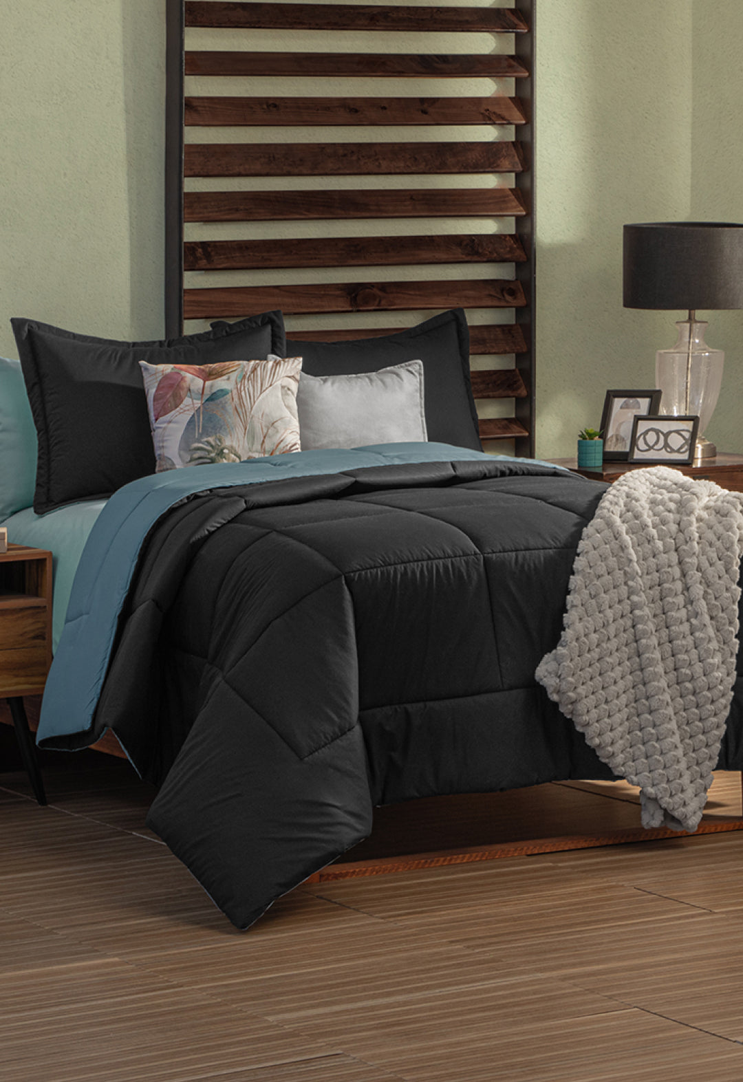 A neatly made bed features the Juego de Edredón Marco by Intima Hogar in black and teal, with decorative pillows, a textured gray throw, and a wooden headboard. A nightstand with a lamp, clock, and decor sits beside the bed.