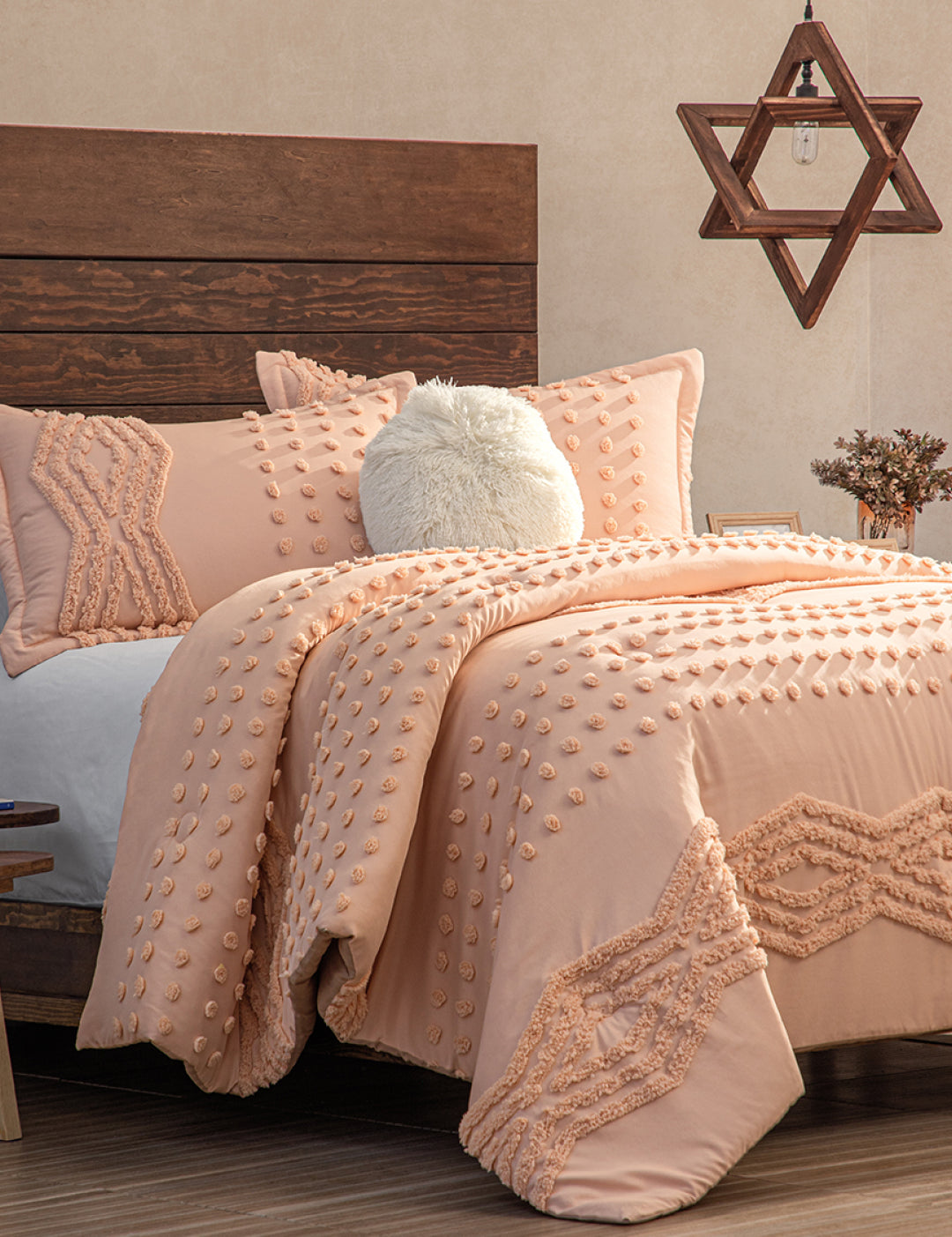 A cozy bedroom features a wooden headboard, the Intima Hogar Juego de Edredón Sedona in peach with textured patterns, a fluffy white pillow, a star-shaped geometric hanging light, neutral walls, and a wooden floor.