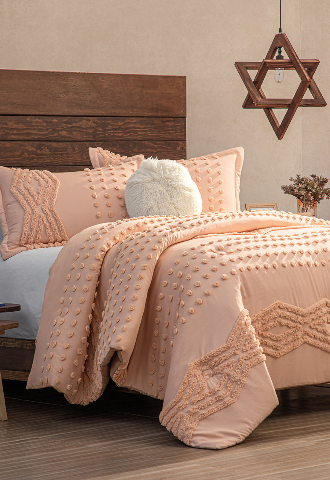 A cozy bedroom features a wooden headboard, the Intima Hogar Juego de Edredón Sedona in peach with textured patterns, a fluffy white pillow, a star-shaped geometric hanging light, neutral walls, and a wooden floor.
