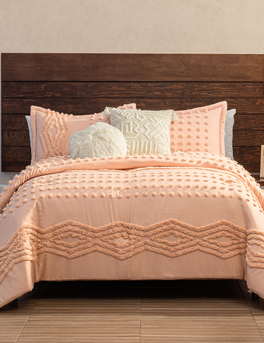 A bed featuring the Intima Hogar Juego de Edredón Sedona in textured peach, with two matching pillows, plus one round and one patterned decorative pillow, set against a dark wooden headboard and beige wall.