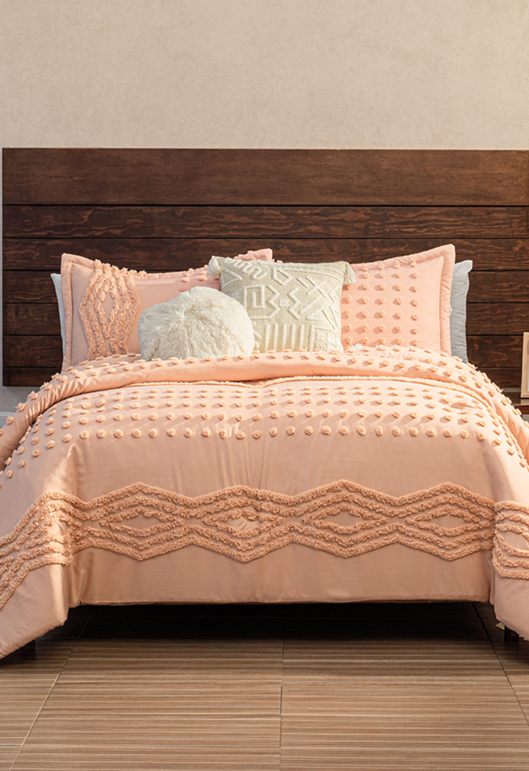 A bed featuring the Intima Hogar Juego de Edredón Sedona in textured peach, with two matching pillows, plus one round and one patterned decorative pillow, set against a dark wooden headboard and beige wall.