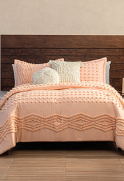 A bed featuring the Intima Hogar Juego de Edredón Sedona in textured peach, with two matching pillows, plus one round and one patterned decorative pillow, set against a dark wooden headboard and beige wall.
