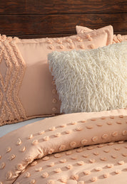 A close-up shows a neatly made bed with the Juego de Edredón Sedona by Intima Hogar in peach, featuring tufted dot patterns on the comforter and pillow, paired with a white shag pillow, set against a wooden headboard.