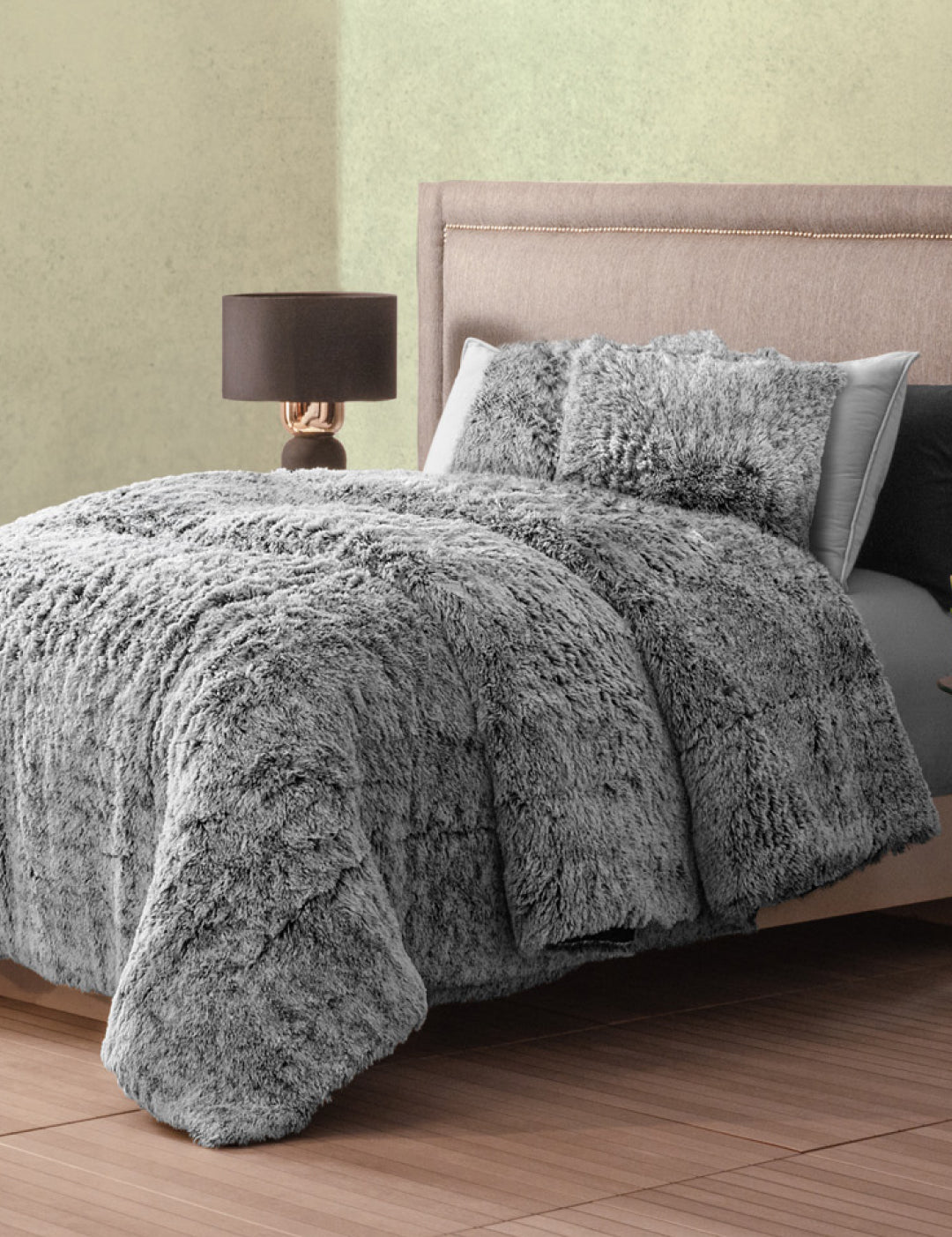 A neatly made bed with Intima Hogar's Juego de Cojines de Lujo Humo and a fluffy gray faux fur comforter rests against a light wall; beside it, a modern nightstand with lamp stands on the wooden floor.