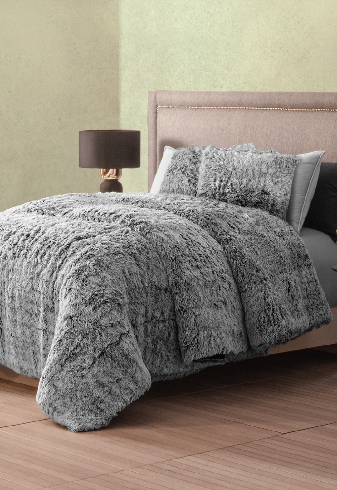 A neatly made bed with Intima Hogar's Juego de Cojines de Lujo Humo and a fluffy gray faux fur comforter rests against a light wall; beside it, a modern nightstand with lamp stands on the wooden floor.