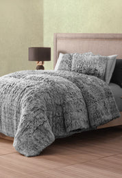 A neatly made bed with Intima Hogar's Juego de Cojines de Lujo Humo and a fluffy gray faux fur comforter rests against a light wall; beside it, a modern nightstand with lamp stands on the wooden floor.