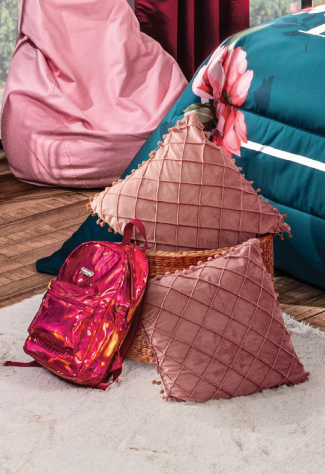 A shiny pink backpack sits on a white rug beside a wicker basket holding two Cojín de Lujo Velvet Rosa by Intima Hogar, in a cozy bedroom with a floral comforter and bean bag chair.