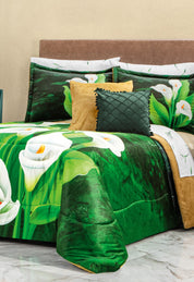 A neatly made bed features bedding with a vivid green and white calla lily design, gold and dark green accent pillows, and the Intima Hogar Cojín de Lujo Velvet Verde Esmeralda. The headboard is upholstered in light brown fabric.