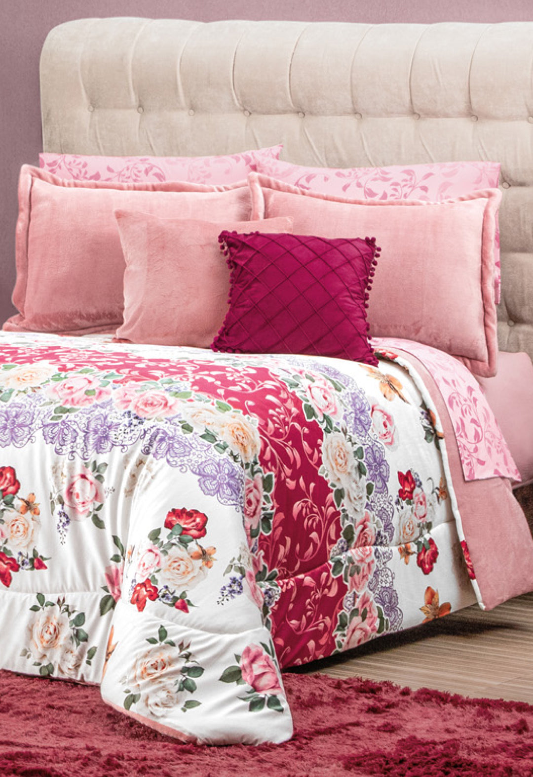 A bed with a beige tufted headboard is decorated with pink and fuchsia pillows, a floral comforter in red, pink, and purple tones, and the Cojín de Lujo Velvet Vino by Intima Hogar adds subtle luxury to the room's modern atmosphere.