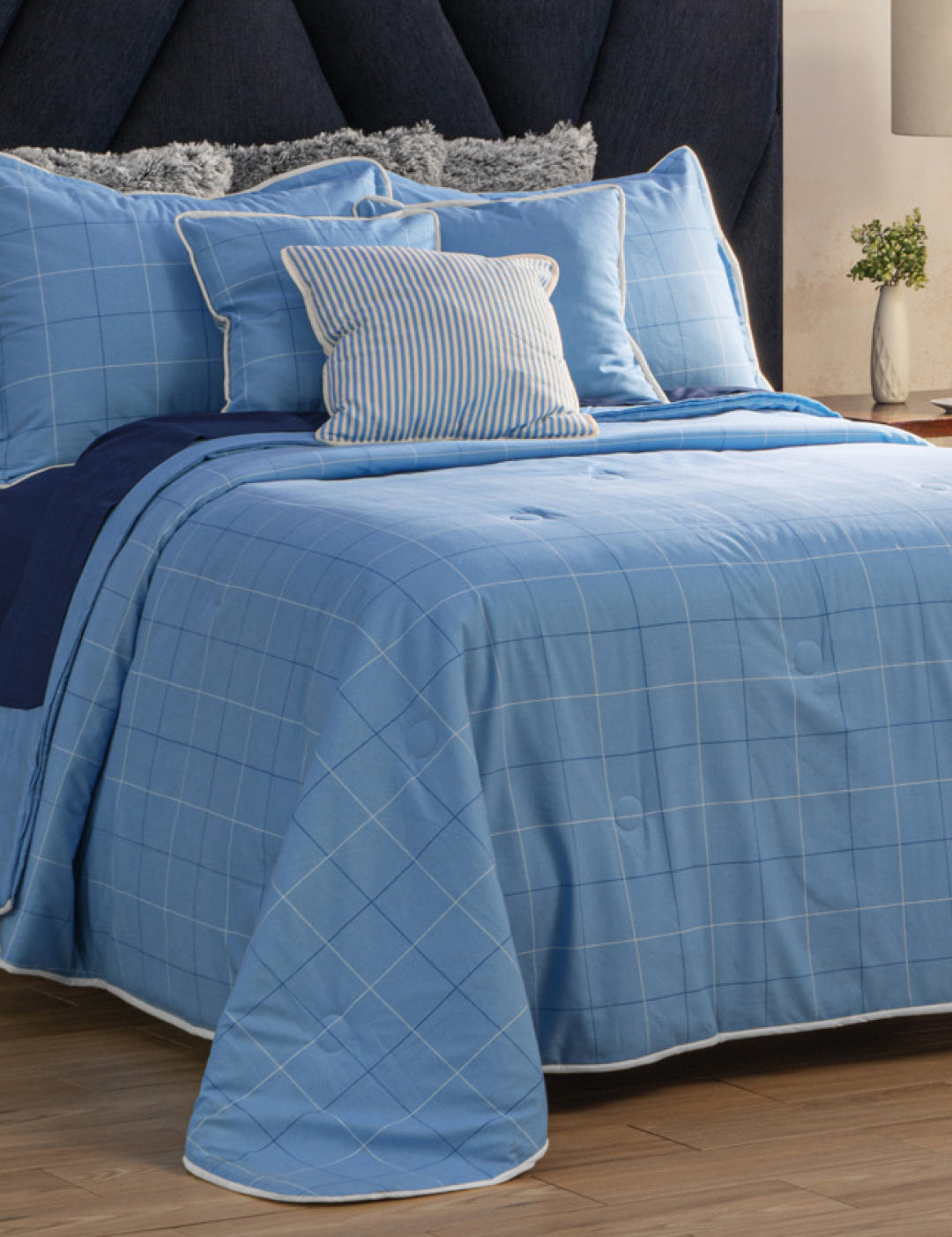 A neatly made bed with the Juego de Edredón Royal Cotton Eleganza from Intima Hogar in light blue checks, several pillows including a striped one, a dark blue headboard, and a nightstand with a plant and lamp in the background.