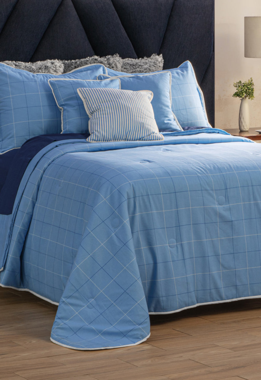 A neatly made bed with the Juego de Edredón Royal Cotton Eleganza from Intima Hogar in light blue checks, several pillows including a striped one, a dark blue headboard, and a nightstand with a plant and lamp in the background.
