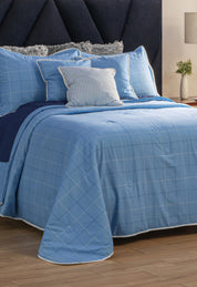 A neatly made bed with the Juego de Edredón Royal Cotton Eleganza from Intima Hogar in light blue checks, several pillows including a striped one, a dark blue headboard, and a nightstand with a plant and lamp in the background.