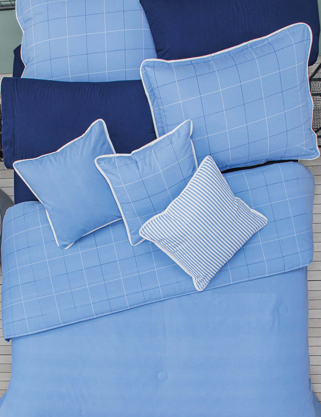 A neatly made bed features the Intima Hogar Juego de Edredón Royal Cotton Eleganza in light and navy blue with solid, grid, and stripe designs. Pillowcases and pillows are arranged in an overlapping, classic style for elegance.