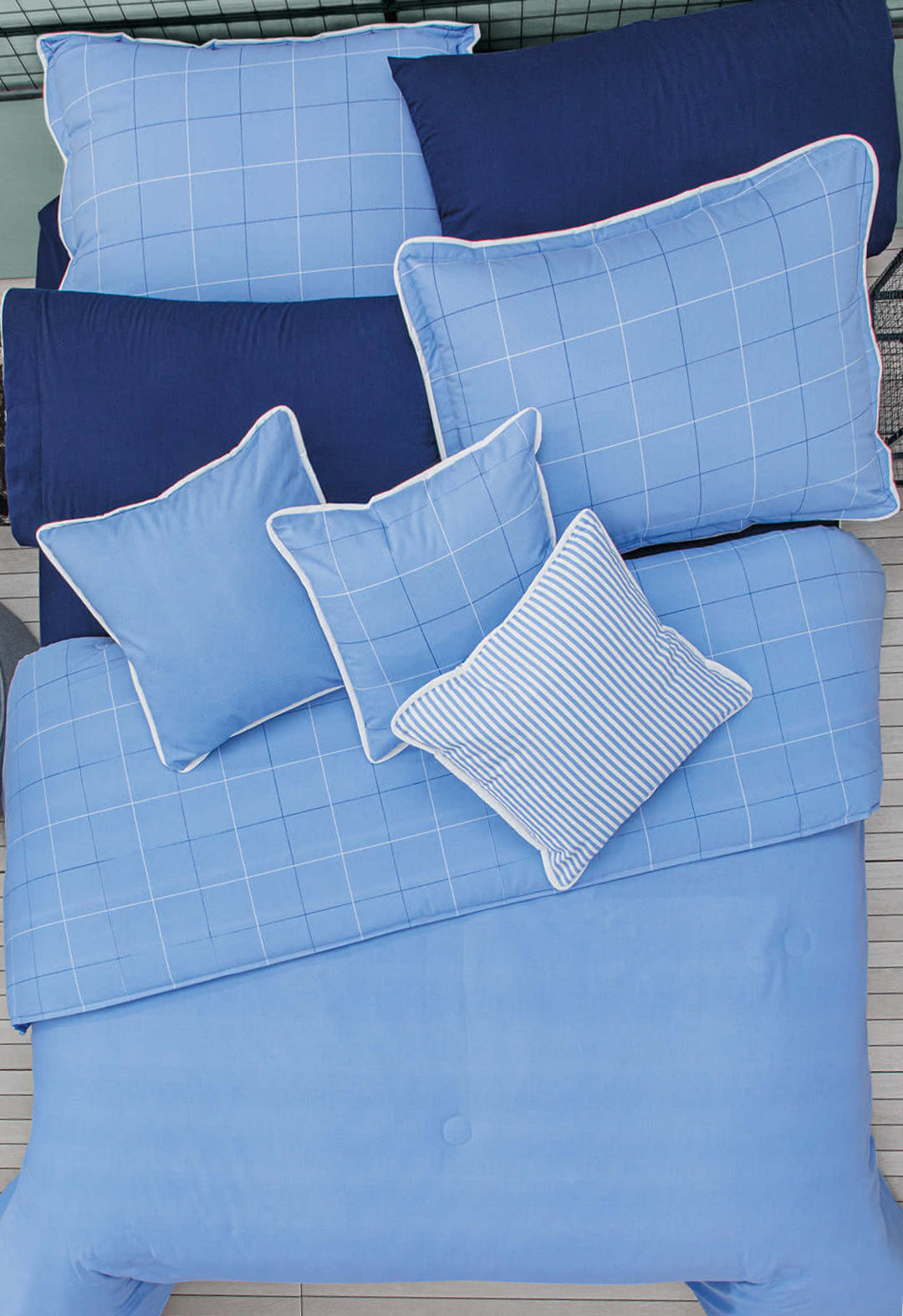 A neatly made bed features the Intima Hogar Juego de Edredón Royal Cotton Eleganza in light and navy blue with solid, grid, and stripe designs. Pillowcases and pillows are arranged in an overlapping, classic style for elegance.