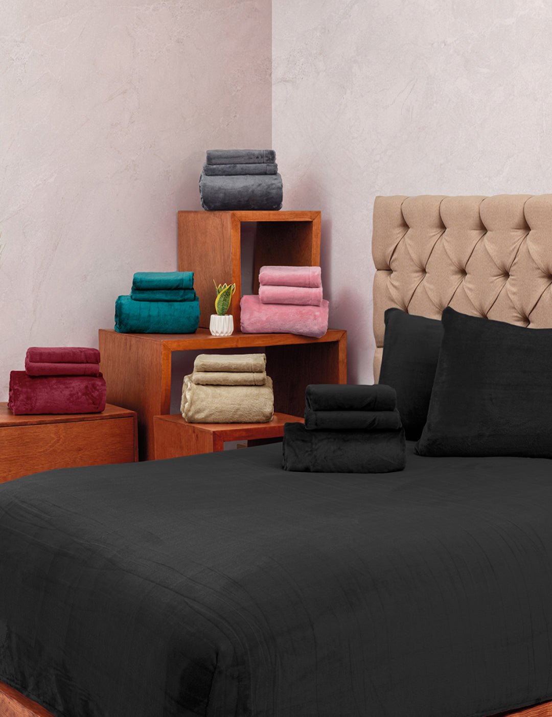 A neatly made bed features the Juego de Sábanas Invernales Negro by Intima Hogar, paired with matching pillows, against a beige tufted headboard. Nearby wooden shelves display towels in teal, red, green, gold, pink, and gray.