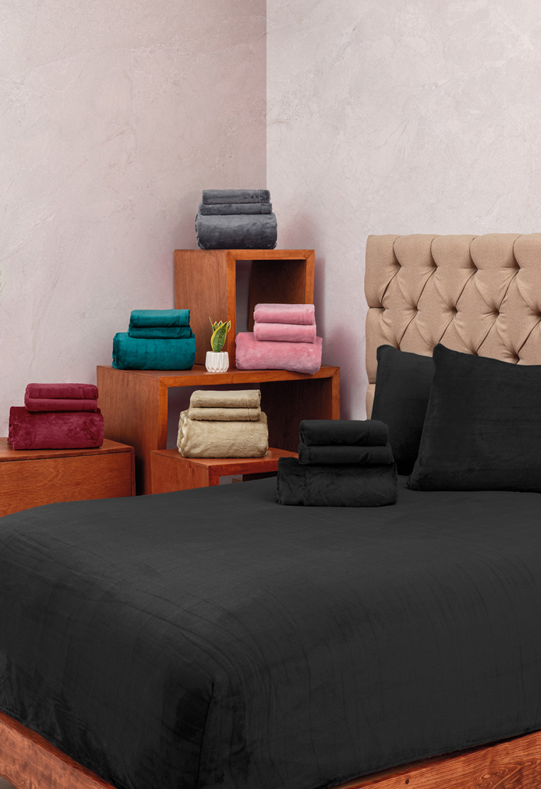 A neatly made bed features the Juego de Sábanas Invernales Negro by Intima Hogar, paired with matching pillows, against a beige tufted headboard. Nearby wooden shelves display towels in teal, red, green, gold, pink, and gray.
