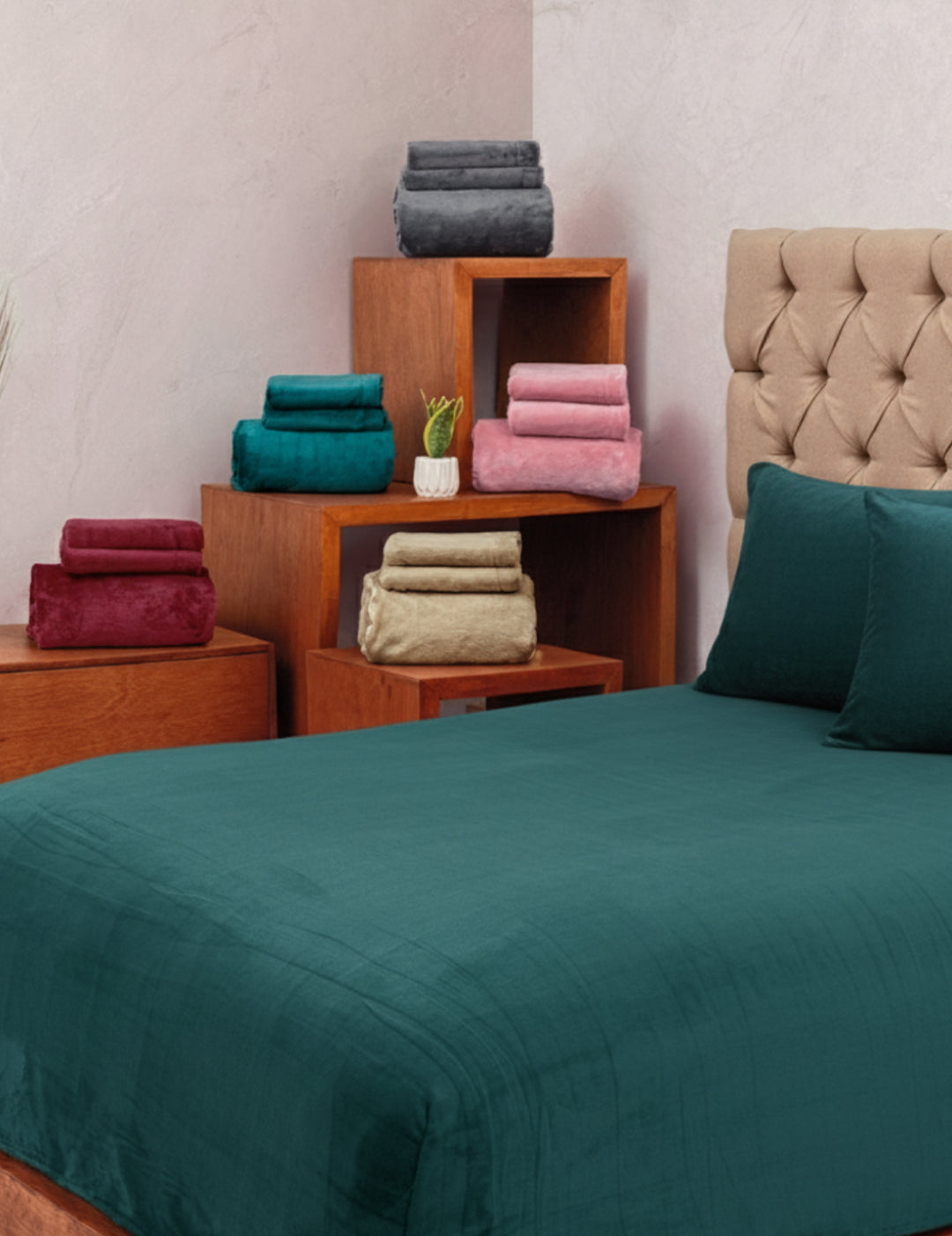 A neatly made bed with Intima Hogar's Juego de Sábanas Invernales Azul Petróleo and matching pillows stands before a beige tufted headboard, while wooden furniture displays folded winter sheets in grey, pink, teal, beige, and red.