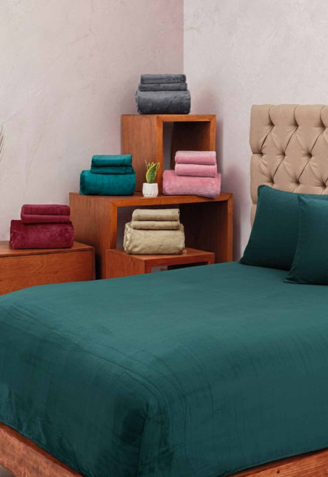 A neatly made bed with Intima Hogar's Juego de Sábanas Invernales Azul Petróleo and matching pillows stands before a beige tufted headboard, while wooden furniture displays folded winter sheets in grey, pink, teal, beige, and red.