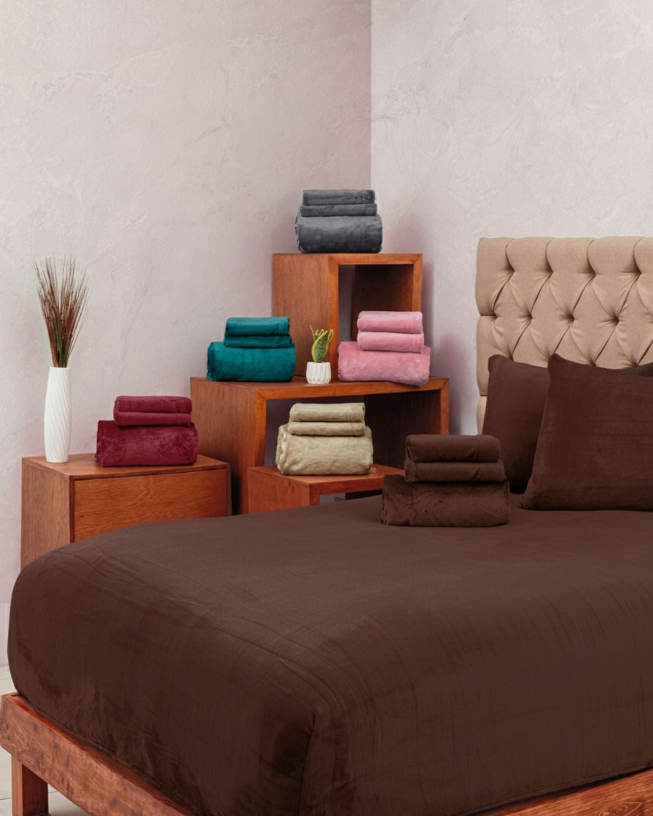 A neatly made bed with the Juego De Sábanas Invernales Café by Intima Hogar and matching pillows, beside wooden nightstands topped with stacks of colorful towels and a white vase with tall grass for decor.