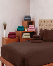 A neatly made bed with the Juego De Sábanas Invernales Café by Intima Hogar and matching pillows, beside wooden nightstands topped with stacks of colorful towels and a white vase with tall grass for decor.