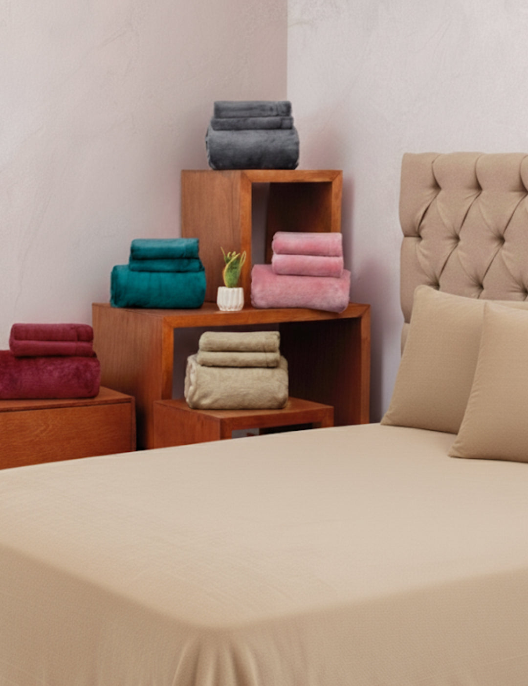 A neatly made bed with the Juego de Sábanas Invernales Camel by Intima Hogar is displayed in front of folded towels in red, green, gray, pink, and beige on wooden shelves and a nightstand beside the bed.