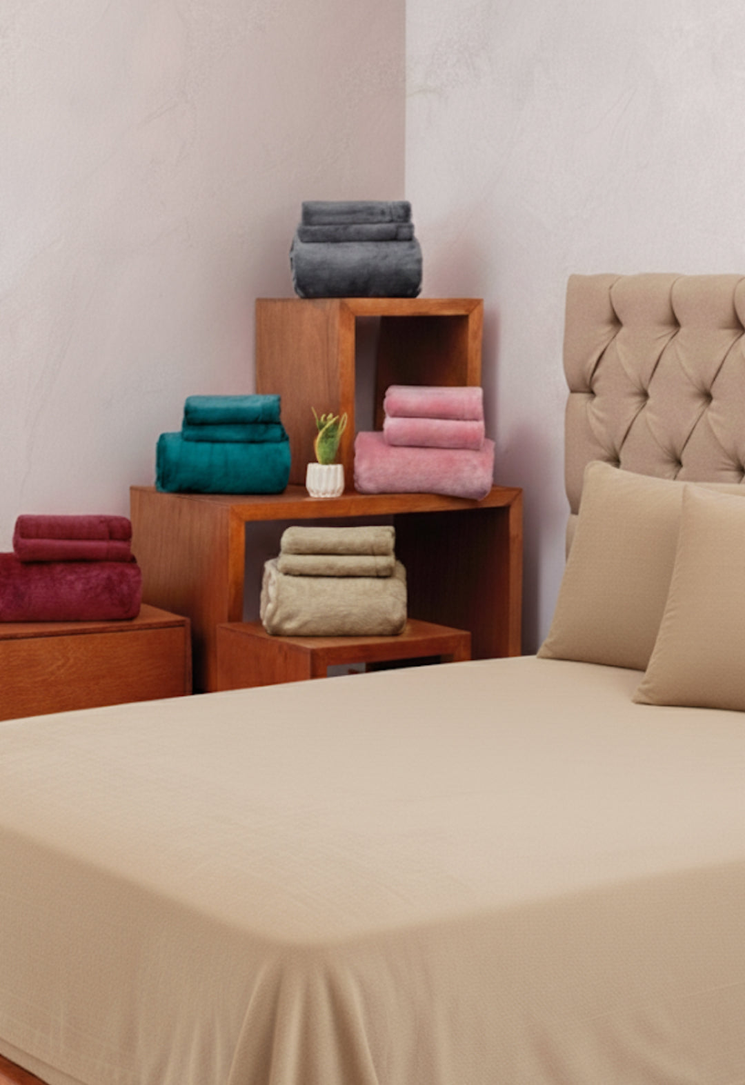 A neatly made bed with the Juego de Sábanas Invernales Camel by Intima Hogar is displayed in front of folded towels in red, green, gray, pink, and beige on wooden shelves and a nightstand beside the bed.