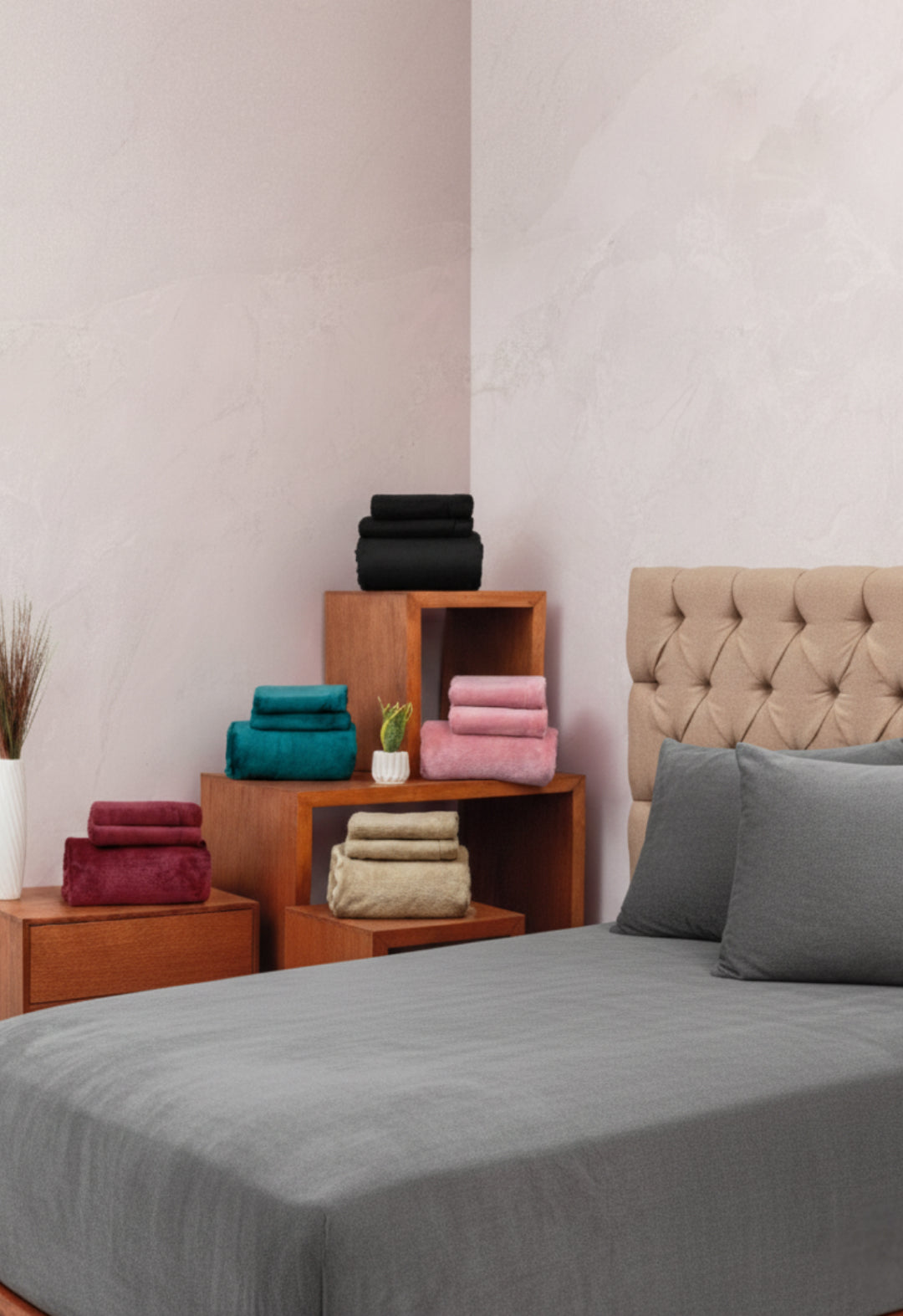 A neatly made bed with the Juego de Sábanas Invernales Gris Oxford by Intima Hogar stands next to wooden shelves stacked with folded towels in teal, burgundy, beige, pink, and black, while a potted plant adds a touch of greenery.