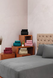 A neatly made bed with the Juego de Sábanas Invernales Gris Oxford by Intima Hogar stands next to wooden shelves stacked with folded towels in teal, burgundy, beige, pink, and black, while a potted plant adds a touch of greenery.