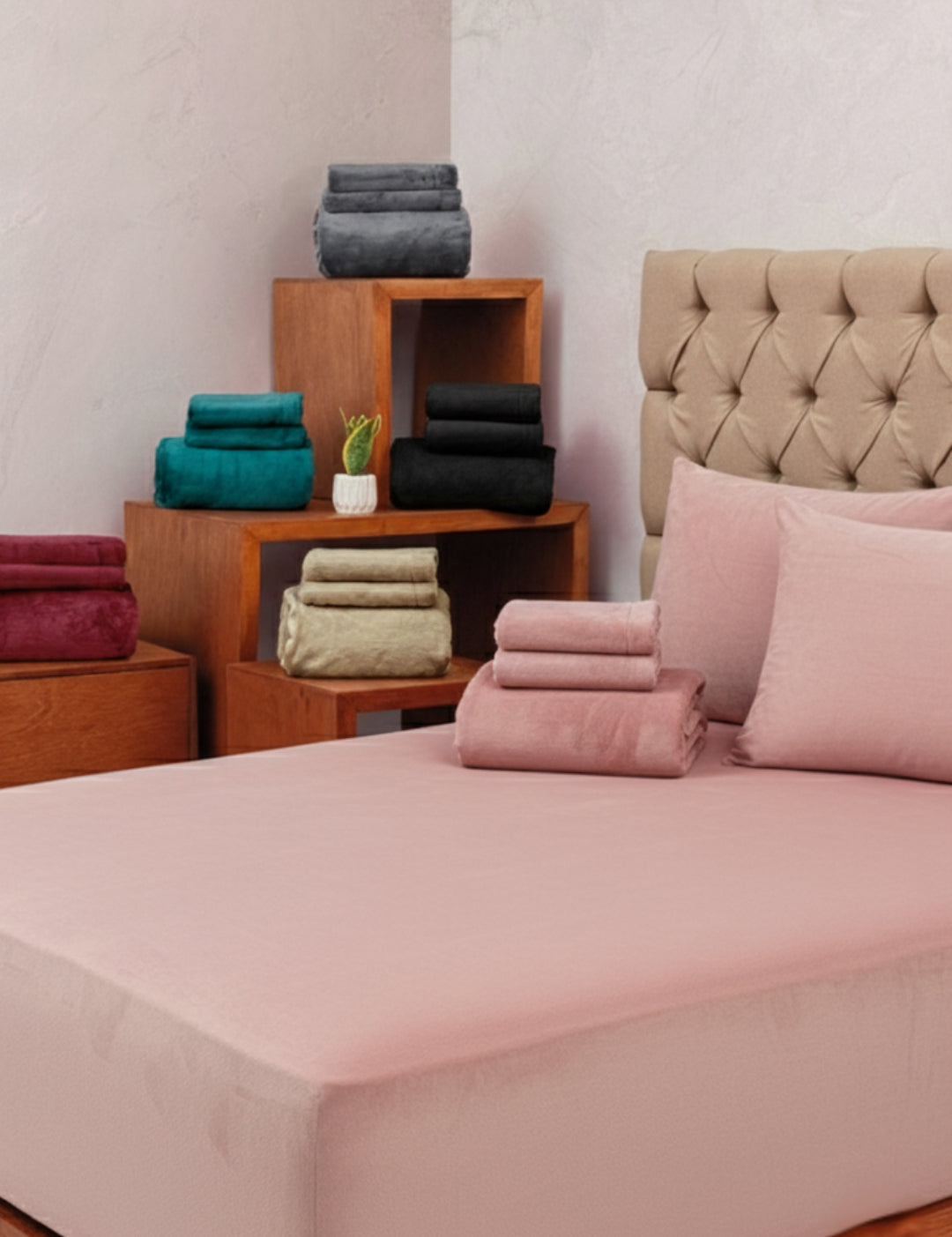 A neatly made bed features the Juego de Sábanas Invernales Palo de Rosa by Intima Hogar with matching pillowcases, stacked with colorful towels. Wooden shelves behind hold more bedding sets and a small potted plant.