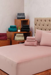 A neatly made bed features the Juego de Sábanas Invernales Palo de Rosa by Intima Hogar with matching pillowcases, stacked with colorful towels. Wooden shelves behind hold more bedding sets and a small potted plant.