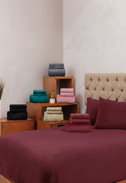 A tufted beige headboard frames a neatly made bed with the Juego de Sábanas Invernales Vino by Intima Hogar, surrounded by folded colorful towels on wooden side tables, set against a light-colored wall.