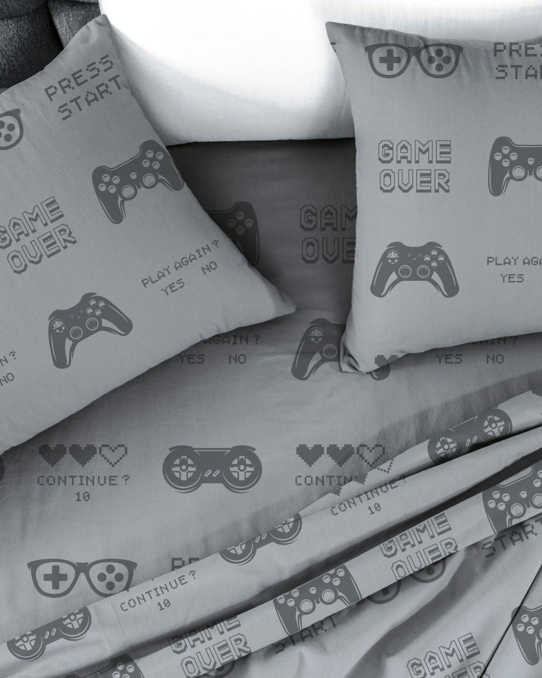 The Juego de Sábanas Play by Intima Hogar features a modern, gamer-themed design with a gray base, video game controller graphics, and fun phrases like GAME OVER, PRESS START, CONTINUE?, plus pixelated hearts. Este set incluye dos fundas de almohada y sábanas perfectas para los fans de los videojuegos.